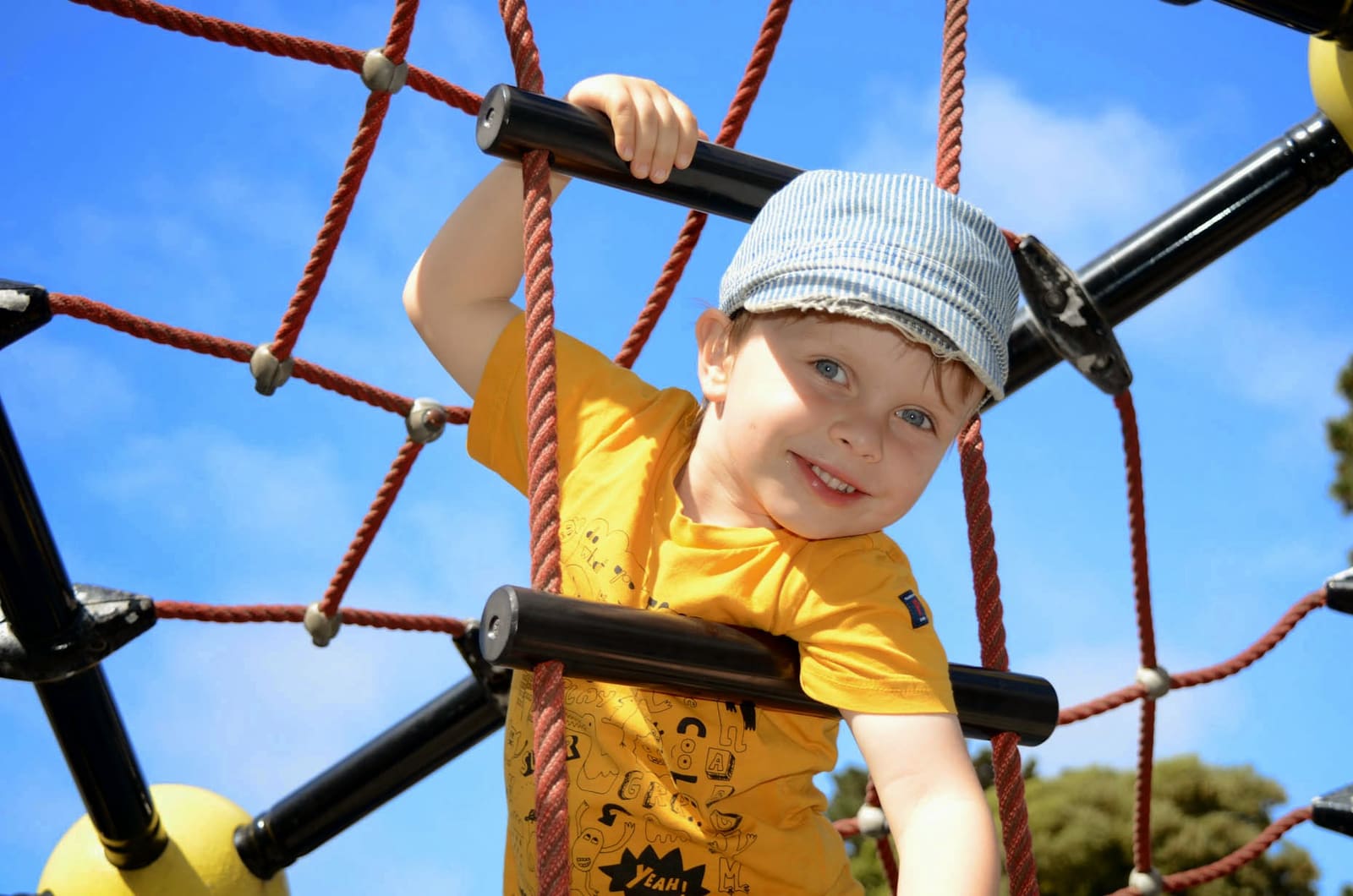 A child in a yellow shirt climbing the rope net of the play structure against a blue sky