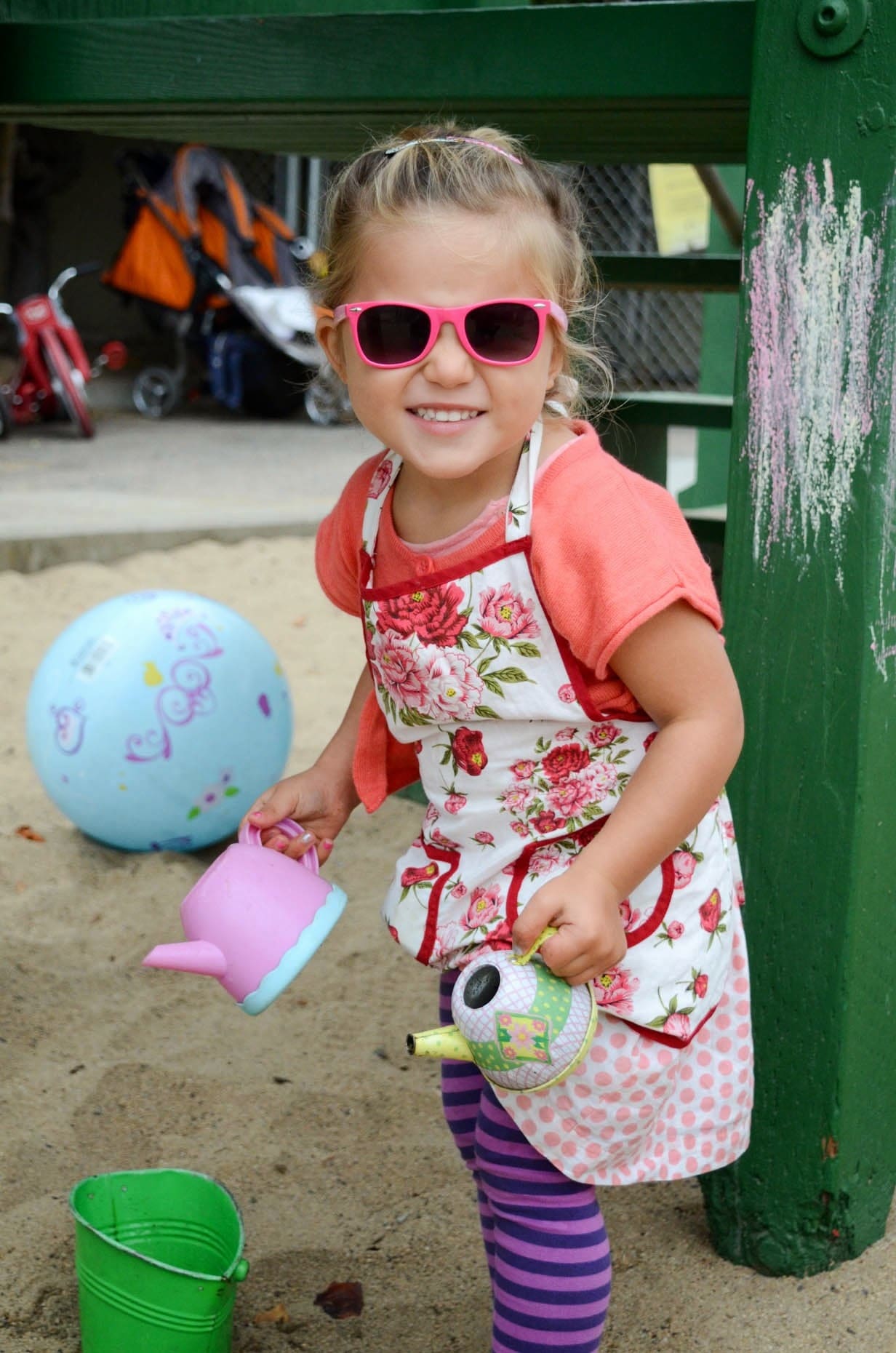 A child in an apron and sunglasses carrying a watering can in the yard