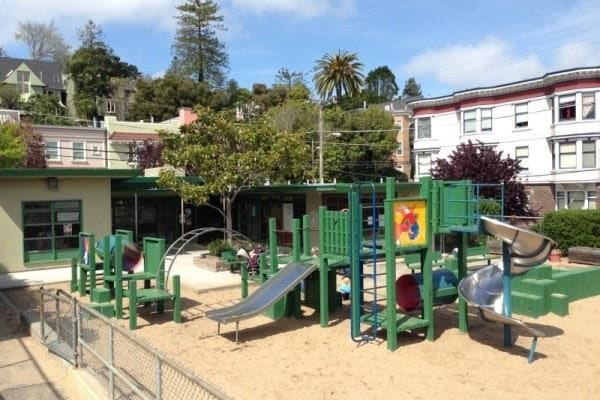 The green play structure and sand yard at Grattan Playground, with Cole Valley houses on the hill behind