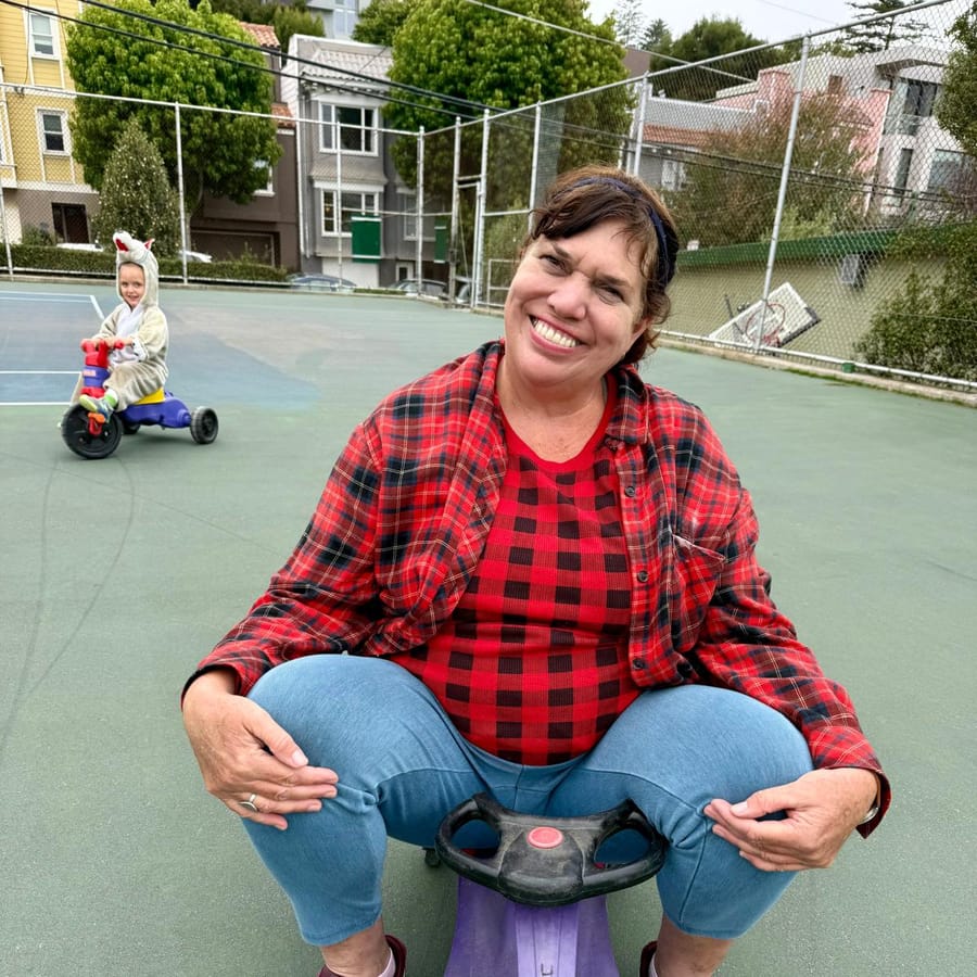 Michele Menard sitting on a child's ride-on toy at the playground, laughing, with a child in a unicorn costume riding a trike behind her
