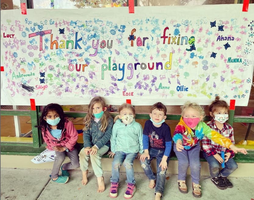 Children sitting beneath a hand-lettered banner reading "Thank you for fixing our playground"